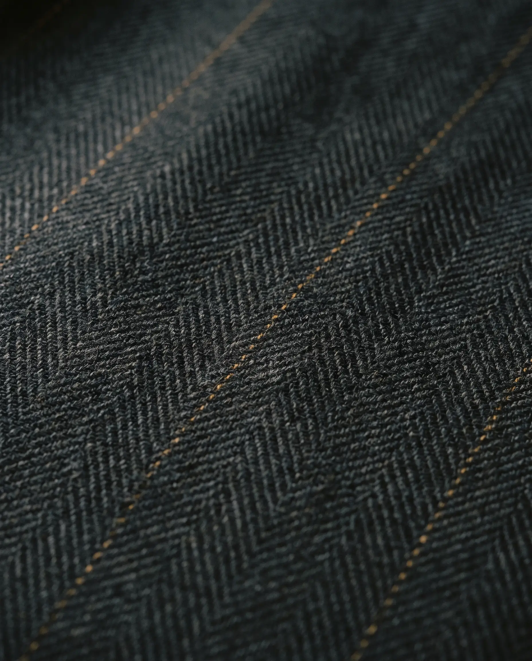 Dark charcoal worsted wool with visible herringbone weave and a subtle antique-gold pinstripe.